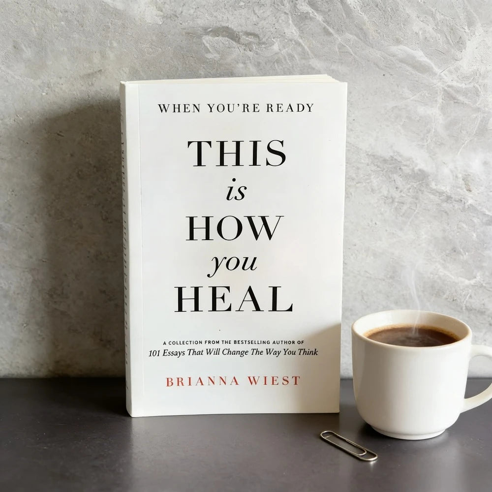 Book titled 'This is How You Heal' by Brianna Wiest with a cup of coffee on a gray surface.