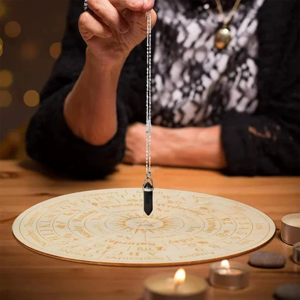 Person holding a pendulum over a circular astrological chart with candles and stones on a wooden surface.