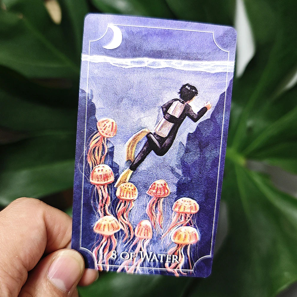 Card from a tarot deck showing a person diving into water with jellyfish, held against a leafy green background.